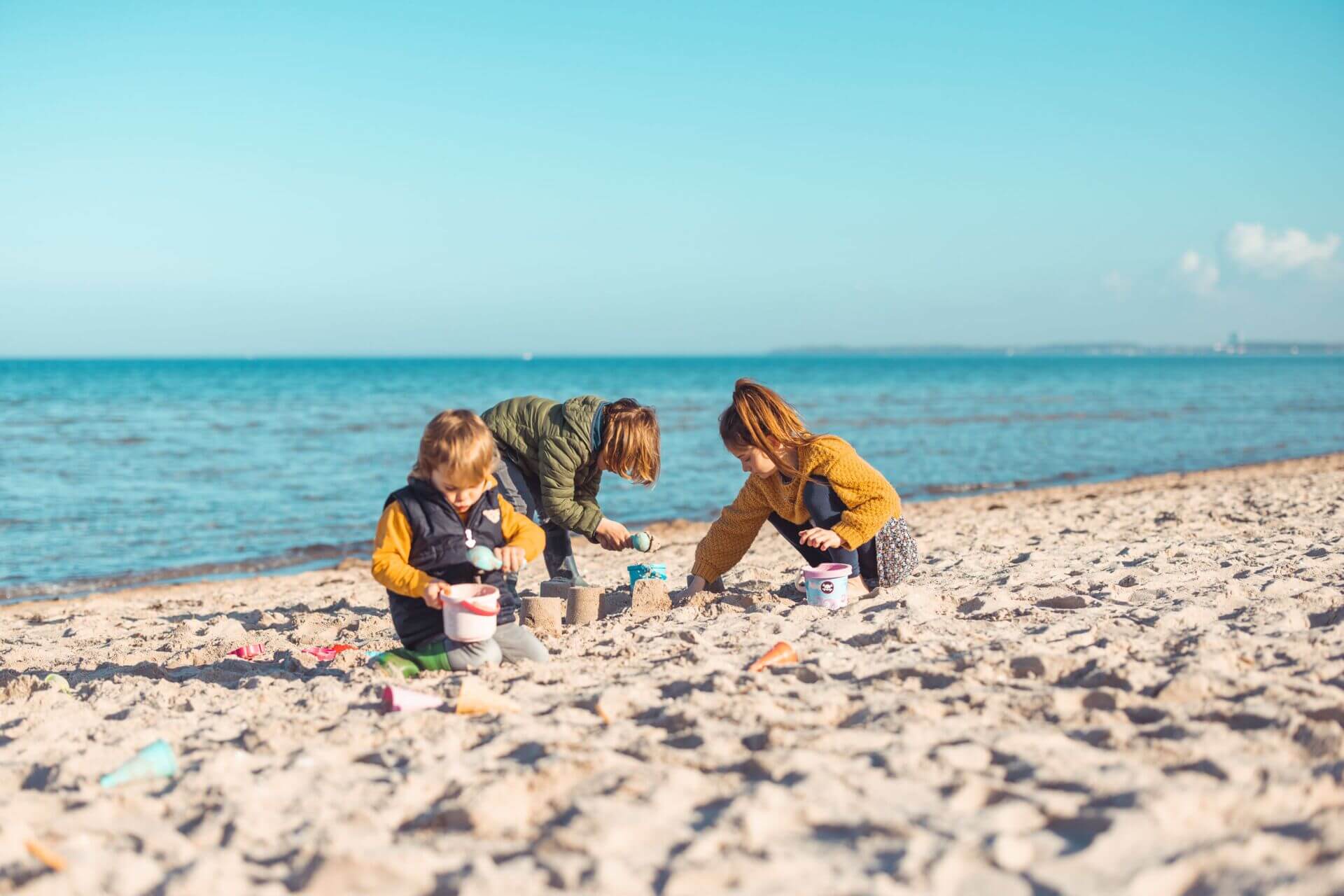 Drei Kinder spielen bei sonnigem Wetter am Ostseestrand im Sand. Das Kind links trägt eine grüne Jacke und ist beschäftigt, Sand in einen Eimer zu füllen, während das mittlere Kind in einer braunen Jacke hilft, eine Sandburg zu bauen. Das Kind rechts im g