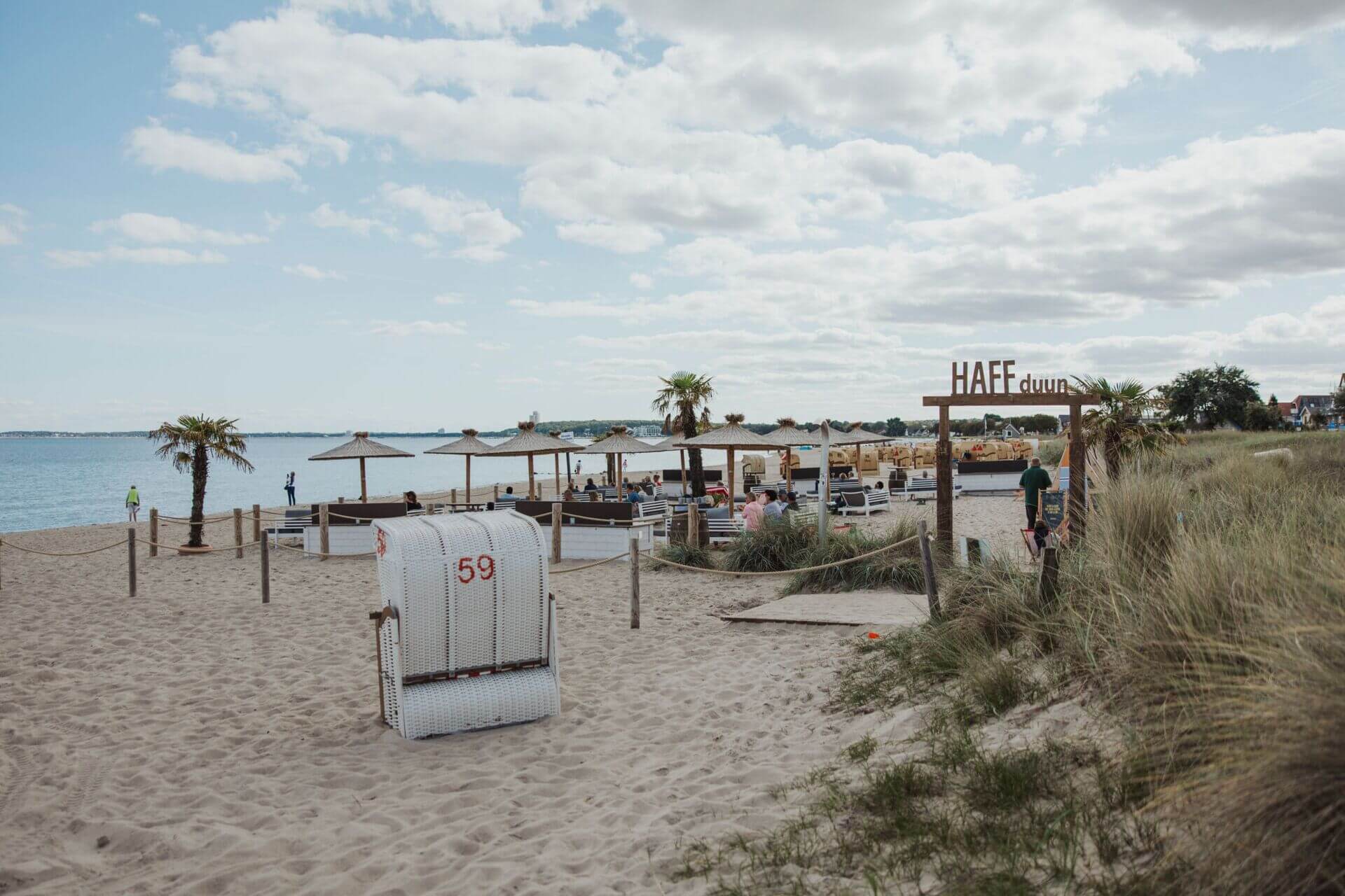 Ein entspannter Strandbereich mit Strandkörben und Sonnenschirmen auf dem Sand, mit Blick auf das Meer. Ein Schild mit der Aufschrift "HAFFdüün" steht an einem Zugangsweg durch das Dünengras. Menschen genießen die Umgebung, einige gehen am Strand entlang,
