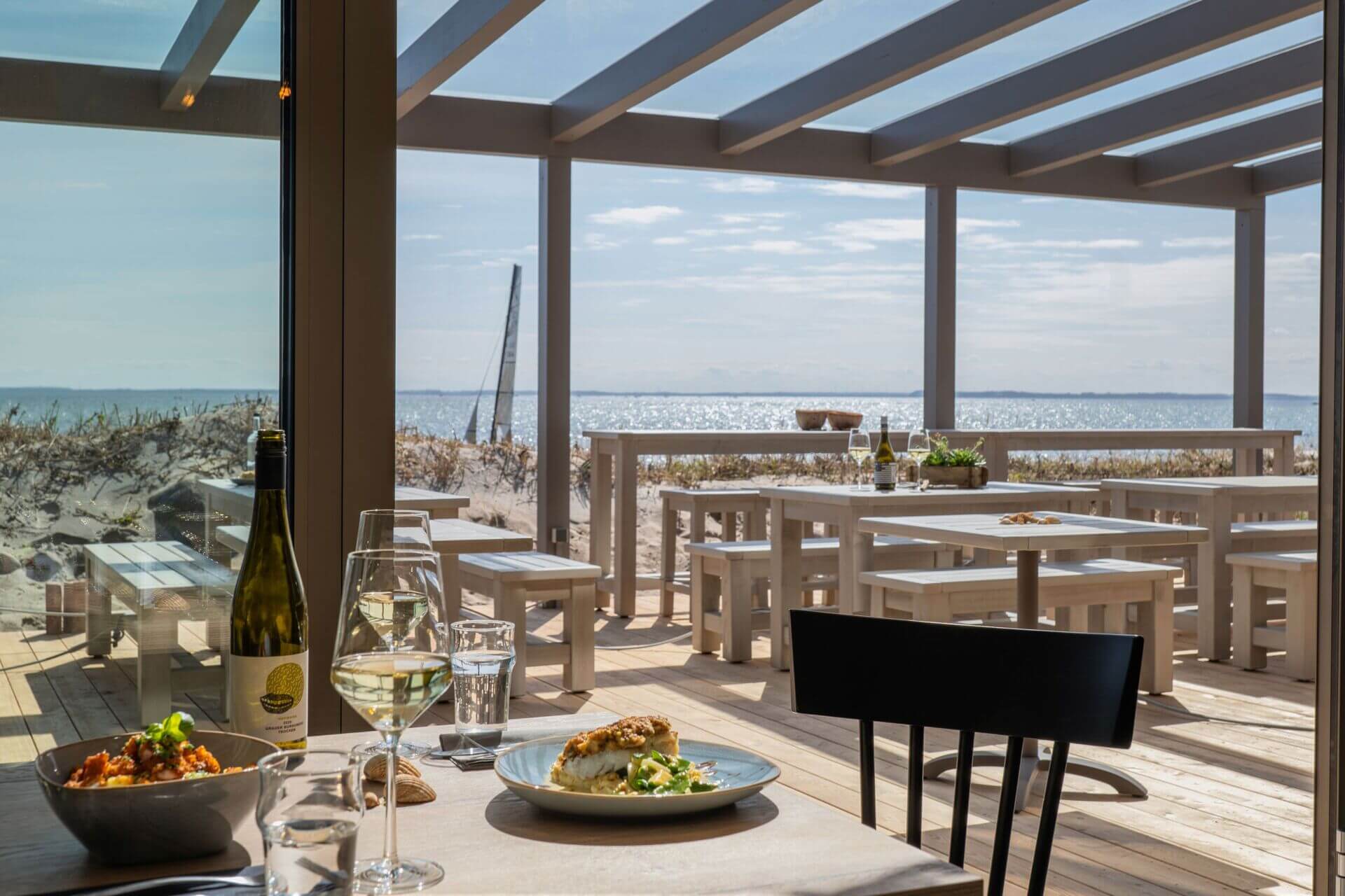 Eine elegante Strandbar mit einer Terrasse bietet einen atemberaubenden Blick auf das Meer. Im Vordergrund steht ein Tisch mit einem Teller Essen, einem Glas Weißwein und einer offenen Flasche Wein. Die Terrasse ist mit modernen, weißen Holzmöbeln ausgest