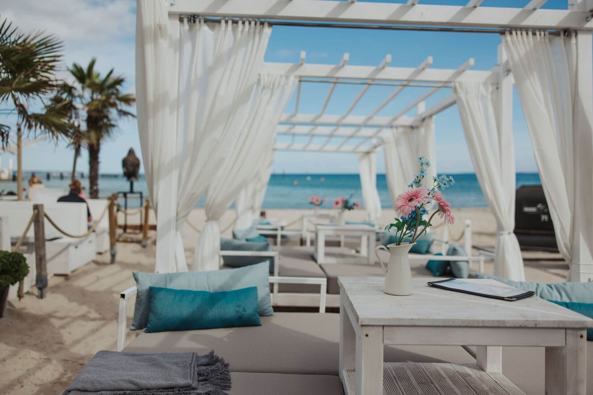 Eine elegante Strandlounge mit bequemen Sitzgelegenheiten, Kissen und einer weißen Pergola und Ostseeblick. Auf einem rustikalen Holztisch steht eine Vase mit rosa Blumen, daneben liegt eine Speisekarte. Im Hintergrund ist der sandige Strand mit dem blaue
