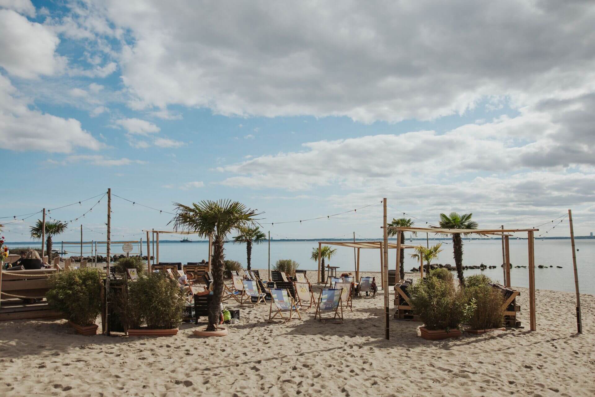 Eine Strandbar direkt am Sandstrand mit einer Reihe von Liegestühlen und rustikalen Holzmöbeln. Ein paar Palmen und große Pflanzenkübel sorgen für ein tropisches Flair. Lichterketten sind oberhalb des Bereichs gespannt, was auf eine gemütliche Atmosphäre